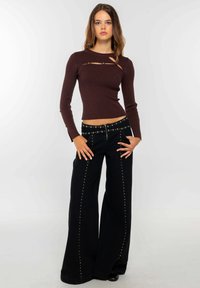 Top marrón oscuro acanalado de manga larga con detalle de corte y botones con acento dorado, combinado con pantalones negros holgados con tachuelas doradas a lo largo de las costuras.