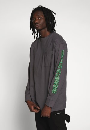 Camiseta de manga larga gris con letras verdes en la manga izquierda, que cuenta con un bolsillo delantero y un corte relajado. Se lleva con pantalones negros.
