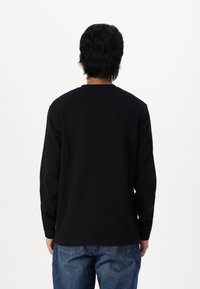 Maglietta nera a maniche lunghe con scollatura a girocollo, realizzata in un tessuto texture. Indossata con jeans chiari a vestibilità ampia, che mostrano un design semplice.