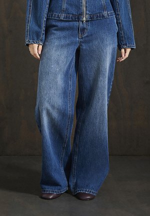 Inimene kannab laia säärega siniseid denimpükse ja sobivat denimpluusi, seistes tumedal põrandal tuhmist taustast.