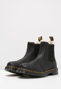 Bottes bottines en cuir noir avec panneaux latéraux élastiques, doublure en fourrure blanche et détails de couture jaune sur la semelle. Semelle extérieure en caoutchouc rainurée.