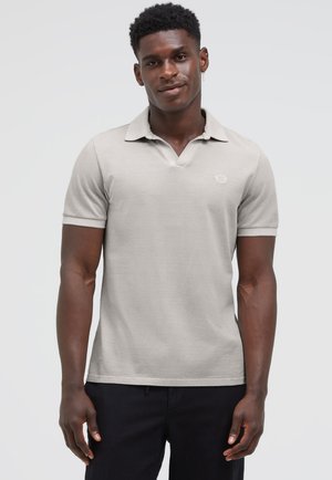 Young man wearing light beige short-sleeve polo shirt and black pants, standing against plain light gray background.