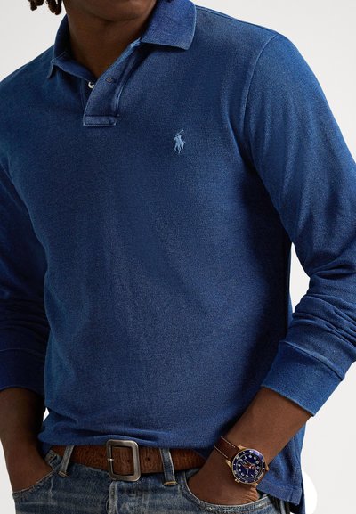Μακρυμάνικο πόλο μπλε ναυτικό με υφή, που φέρει μπλε κεντημένο λογότυπο και φοριέται με τζιν και καφέ ζώνη.
