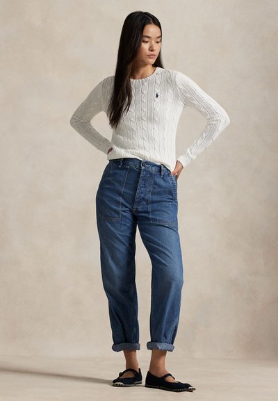 Vrouw met lang donker haar die een witte kabeltrui, opgerolde blauwe jeans en zwarte platte schoenen draagt, staat met haar handen in haar zij.