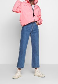 Neon růžový větrový kabát se zipovým zapínáním, vysokým límcem a elastickými manžetami, kombinovaný s modrými džínami rovného střihu a béžovými vysokými teniskami.