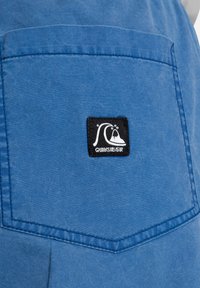 Plava tkanina džepa s crno-bijelim Quiksilver logotipom, sa šavovima oko rubova i glatkom teksturom.
