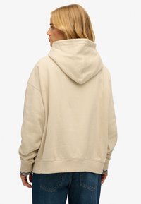 Beżowa bluza z dużym kapturem, luźnym krojem i ściągaczami na rękawach, zestawiona z niebieskimi dżinsami. Tkanina wydaje się miękka o gładkiej fakturze.