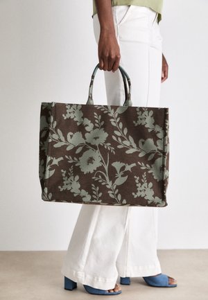Personne tenant un grand sac cabas marron avec des motifs floraux verts, portant un pantalon blanc et des chaussures bleues à talons blocs et à bouts ouverts.