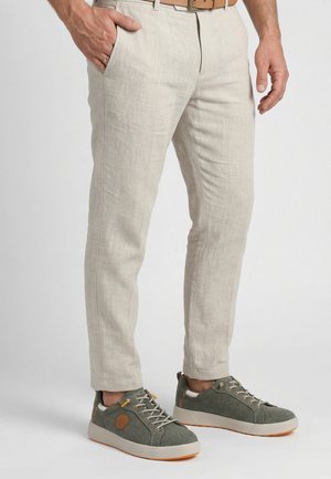 Muž v béžových kalhotách s jednou rukou v kapse a zelených sportovních teniskách s bílou podrážkou a oranžovými akcenty.