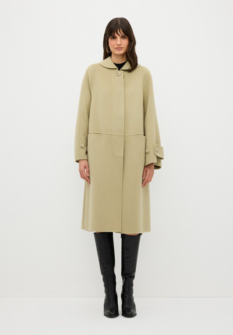 Cappotto di lana beige con collo alto, bottoni sulle maniche e due tasche frontali. Abbinato a stivali neri al ginocchio.