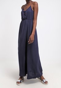 Marineblauwe maxidress van lichte stof met een V-hals, geraffineerde taille en verstelbare spaghettibandjes; gecombineerd met metallic sandalen.