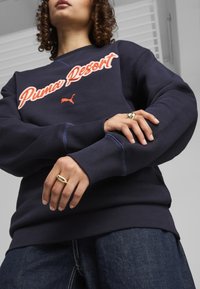 Sudadera azul marino con bordado de "Puma Resort" y logotipo. Presenta puños y dobladillo de canalé, tejido texturizado y costuras contrastantes.