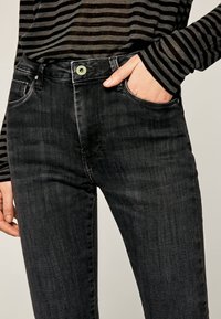 Person som bär svarta tvättade jeans och en långärmad skjorta med svarta och mörkgrå ränder, med ena handen i framfickan.
