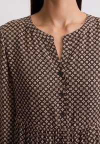 Robe à motifs marron avec un décolleté en V et quatre boutons noirs. Présente un tissu texturé avec un motif floral et des fronces à la taille.
