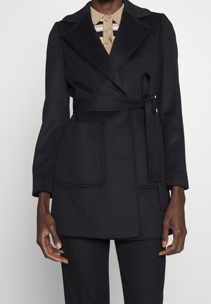 Person iført en sort frakke med bælte over en beige skjorte med krave, knapper og striber, stående foran en ensfarvet baggrund.