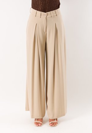 Pantaloni beige a gamba larga con pieghe indossati con sandali bianchi aperti in punta su una persona con un top in pizzo marrone.