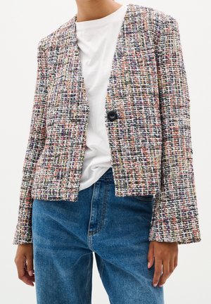 Vrouw die een meerkleurig tweedblazer draagt met een enkele zwarte knoop, een wit overhemd en een blauwe spijkerbroek, staand tegen een effen lichte achtergrond.