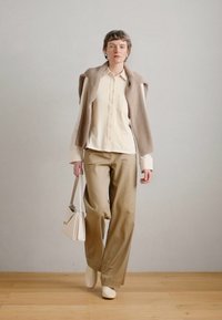 Μπεζ πλεκτό γιλέκο πάνω από κρεμ πουκάμισο με κουμπιά, συνδυασμένο με φαρδιούς καφέ παντελόνες και ταιριαστά κρεμ παπούτσια, κρατώντας μια μικρή λευκή τσάντα.