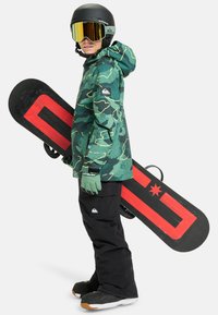 Un snowboarder îmbrăcat într-o jachetă de camuflaj și pantaloni negri ține un snowboard cu un design roșu. Accesoriile includ mănuși și o cască neagră cu ochelari de protecție.