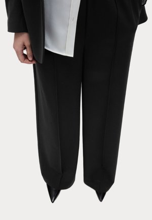 Personne portant un pantalon noir à jambes larges, des chaussures noires pointues, une chemise blanche et un blazer noir avec une main visible sur le côté.