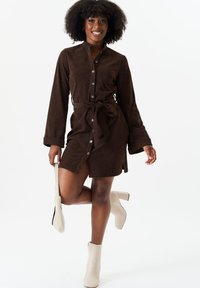 Brown corduroy shirt dress with long sleeves, buttons down the front, tie waist, and side slit. Paired with cream block-heel ankle boots.