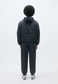 Survêtement léger noir avec capuche, doté d'une poche de dos zippée et de poignets élastiques, associé à des baskets blanches et noires.