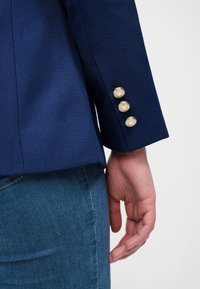 Marineblauwe blazer met een herringbone structuur, voorzien van drie gouden knopen op de manchetten en een slim fit, gecombineerd met blauwe denim.