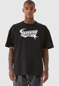 Svart oversized t-skjorte laget av bomull, med et stort hvitt grafisk logo med stilisert tekst og en stjernedesign foran.