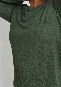 Blusa verde texturizzata con scollo drappeggiato e linee diagonali. Il tessuto sembra morbido e leggermente lucido, con spacco laterale per un tocco di dettaglio in più.