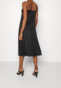 Robe nuisette en satin noir avec fines bretelles, jupe évasée fluide et détails plissés, associée à des sandales à talons carrés noires.