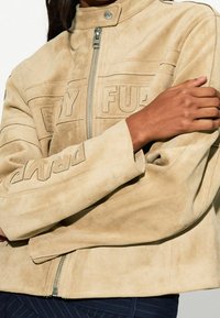 Beżowa zamszowa kurtka z wysokim kołnierzem, przednim zamkiem błyskawicznym i wytłaczanym napisem. Posiada długie rękawy i teksturowane, miękkie wykończenie.