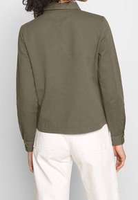 Olivově zelená bunda s límcem, dlouhými rukávy a knoflíkovými manžetami, vyrobená z hladkého materiálu. Má rovný lem a minimalistické detaily šití.