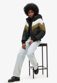Pufferjack met kleurblokken in zwart, olijfgroen en wit; heeft een ritssluiting en capuchon. Model draagt lichtblauwe jeans en zwarte schoenen.