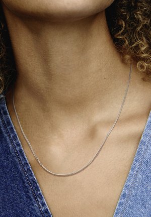 Silberne Halskette mit glatter, glänzender Oberfläche, die flach auf einem nackten Hals liegt und durch ein Denim-Gewand ergänzt wird.