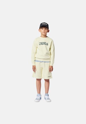 Sweatshirt jaune clair avec le texte "ZADIG&", associé à un short jaune assorti. Casquette noire et baskets blanches avec des accents bleu clair.