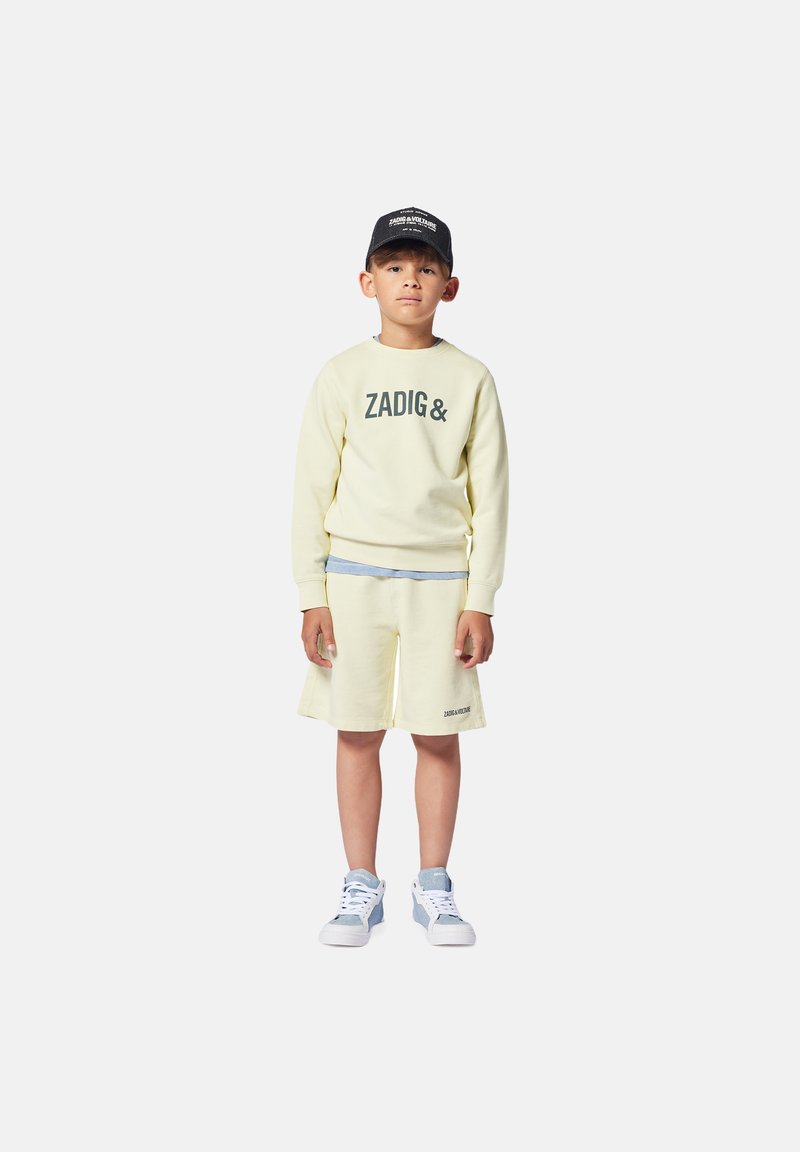 Sweatshirt jaune clair avec le texte "ZADIG&", associé à un short jaune assorti. Casquette noire et baskets blanches avec des accents bleu clair.