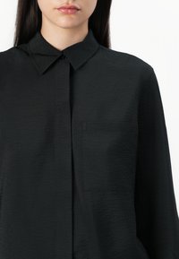 Svart skjorta med knappar, tillverkad av texturerad tyg, med klassisk krage och en enda bröstficka. Enkel, modern design.