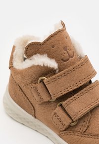 Chaussure montante en cuir marron avec doublure en fourrure, présentant un design de visage d'ours sur la languette et des sangles Velcro ajustables pour la fermeture.