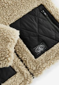 Fleecejacke mit einer weichen beigen Textur, ausgestattet mit einer schwarzen gesteppten Stofftasche und einem Reißverschluss, sowie einem Logoschild an der Tasche.