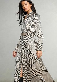 Lungo vestito a camicia in tessuto crema con strisce geometriche e motivi neri. Presenta un colletto e una vita accentuata da una cintura per definire la silhouette.