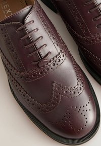 Chaussures habillées en cuir bordeaux avec un design à perforations et à bouts rabats, fermeture à lacets et semelle en caoutchouc texturée. Des coutures détaillées soulignent la tige.