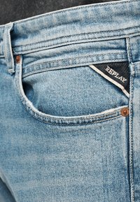 Jean en denim bleu clair en gros plan montrant la poche arrière avec une étiquette noire portant l'inscription "REPLAY" et des rivets en cuivre sur les bords de la poche.