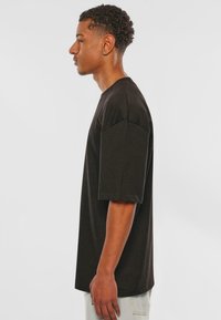Schwarzes T-Shirt mit kurzen Ärmeln, aus Baumwollstoff, lockerer Schnitt, runder Ausschnitt, glatte Textur, mit dezenten Seitenlogo-Details, kombiniert mit hellen Hosen.