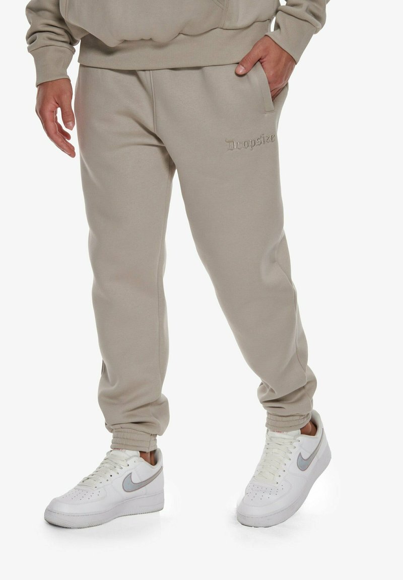 Beige tepláky s volným střihem, zahrnující boční kapsy a žebrované manžety. Na přední straně je embosované logo "Dropsize". V kombinaci s bílými teniskami.