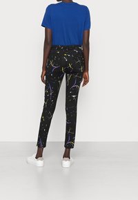 Persona vistiendo pantalones negros ajustados con salpicaduras de pintura abstracta coloridas, camiseta de manga corta azul y zapatillas blancas, de pie de espaldas.