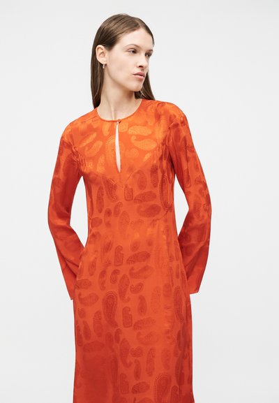 Γυναίκα που φοράει πορτοκαλί φόρεμα με μακριά μανίκια, διακοσμημένο με διακριτικά σχέδια παϊέτας και μικρή σχισμή μπροστά, στέκεται μπροστά σε απλό φόντο.