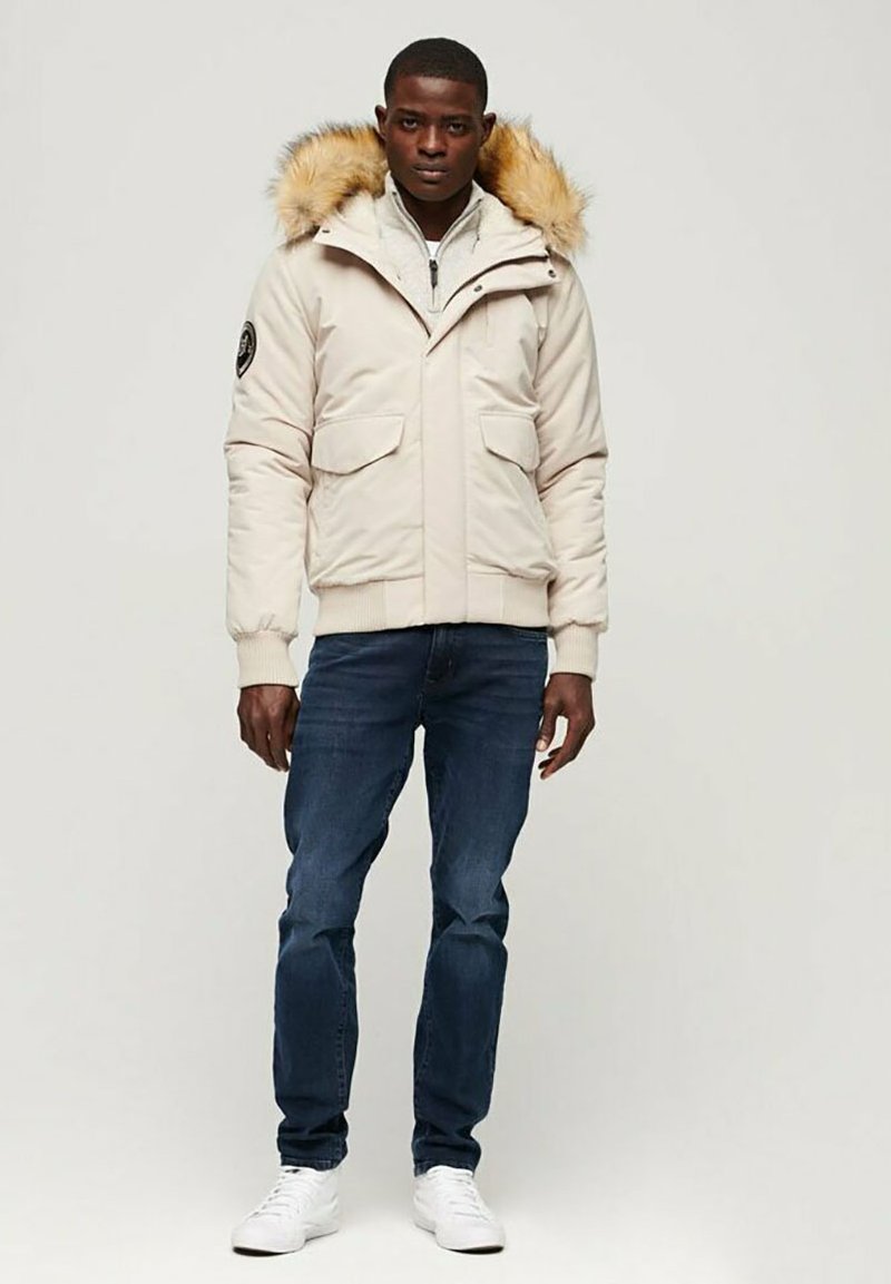 Helle beige Puffjacke mit einer mit Fell gefütterten Kapuze, Reißverschluss vorne, zwei Brusttaschen und gerippten Bündchen. Getragen über einem grauen Reißverschlussoberteil und blauen Jeans.