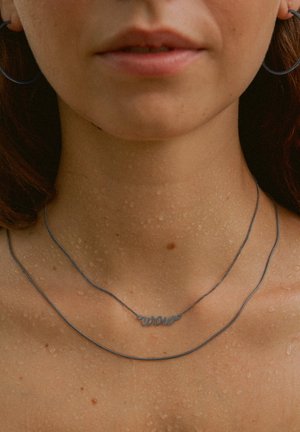 Silberne Halskette mit "Wow"-Anhänger, die ein glattes, elegantes Design aufweist; kombiniert mit einer dünnen Kette. Wassertropfen auf der Haut verleihen Textur.