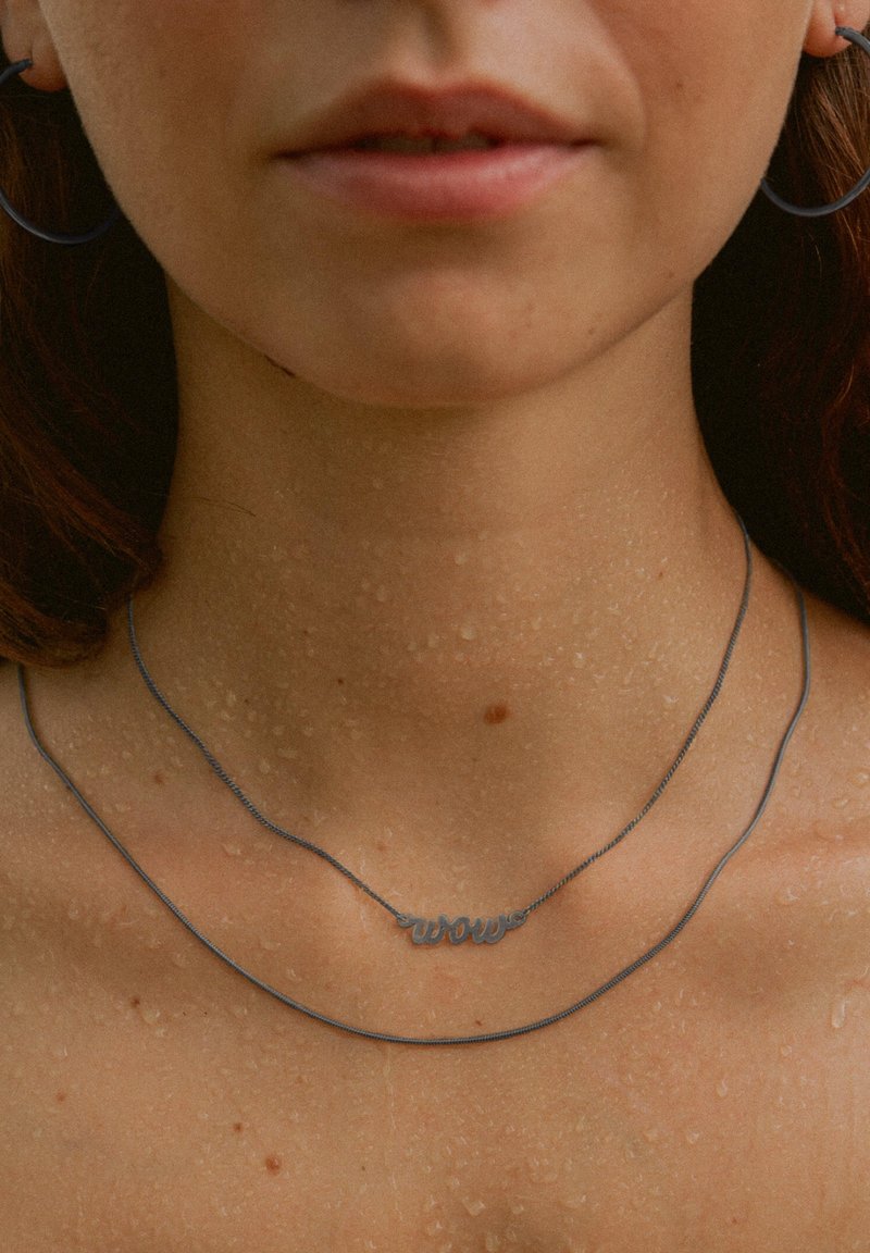 Silberne Halskette mit "Wow"-Anhänger, die ein glattes, elegantes Design aufweist; kombiniert mit einer dünnen Kette. Wassertropfen auf der Haut verleihen Textur.