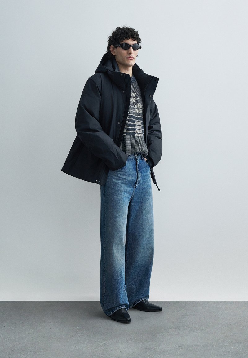 Black waterproof jacket, gray patterned sweater, light blue wide-leg jeans, and black pointed shoes. Model wears dark sunglasses.